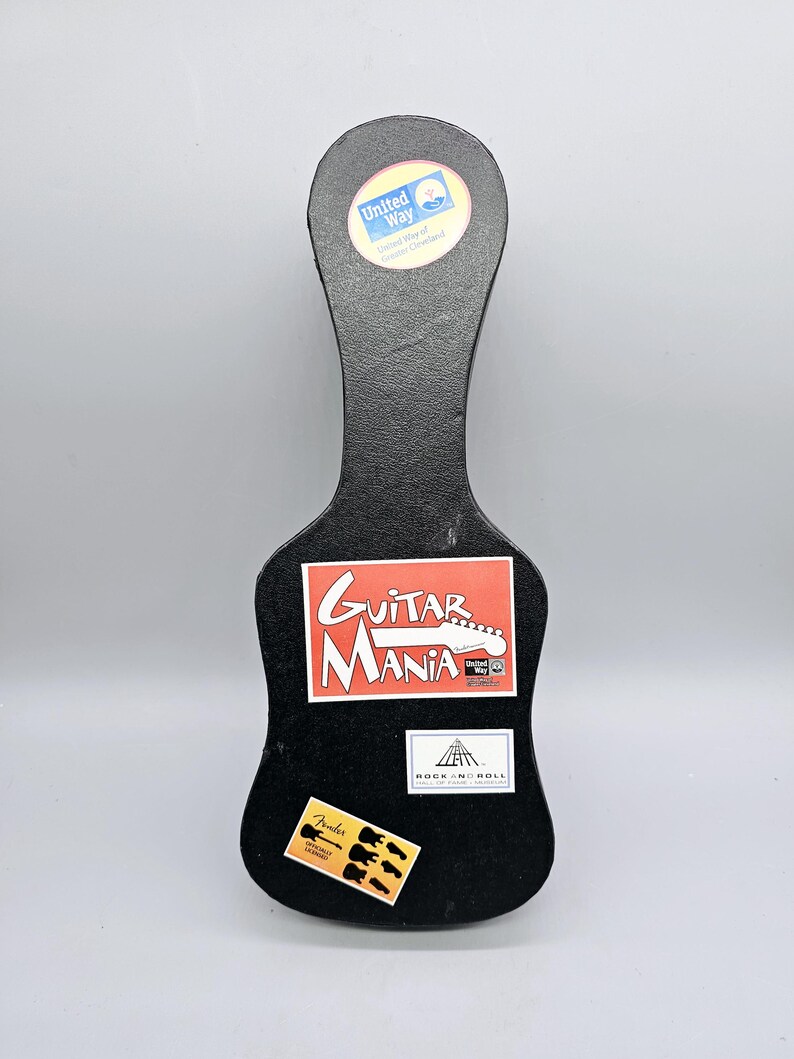 May include: Black guitar-shaped box with stickers. The stickers include "Guitar Mania", "Rock 'n' Roll", "Fender", and a United Way logo.
