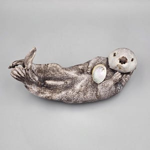 May include: A ceramic sea otter figurine with a textured gray and brown mottled finish. The otter is lying on its back, holding a small, iridescent, oval-shaped abalone shell against its chest with its front paws, while its tail is curled.