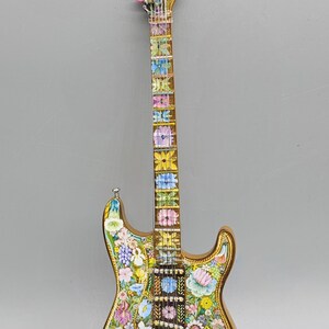 May include: A gold electric guitar with a floral design. The guitar has a pink flower on the headstock and the words "Fender Stratocaster" are visible. The body of the guitar is covered in a variety of flowers, including roses, daisies, and lilies.