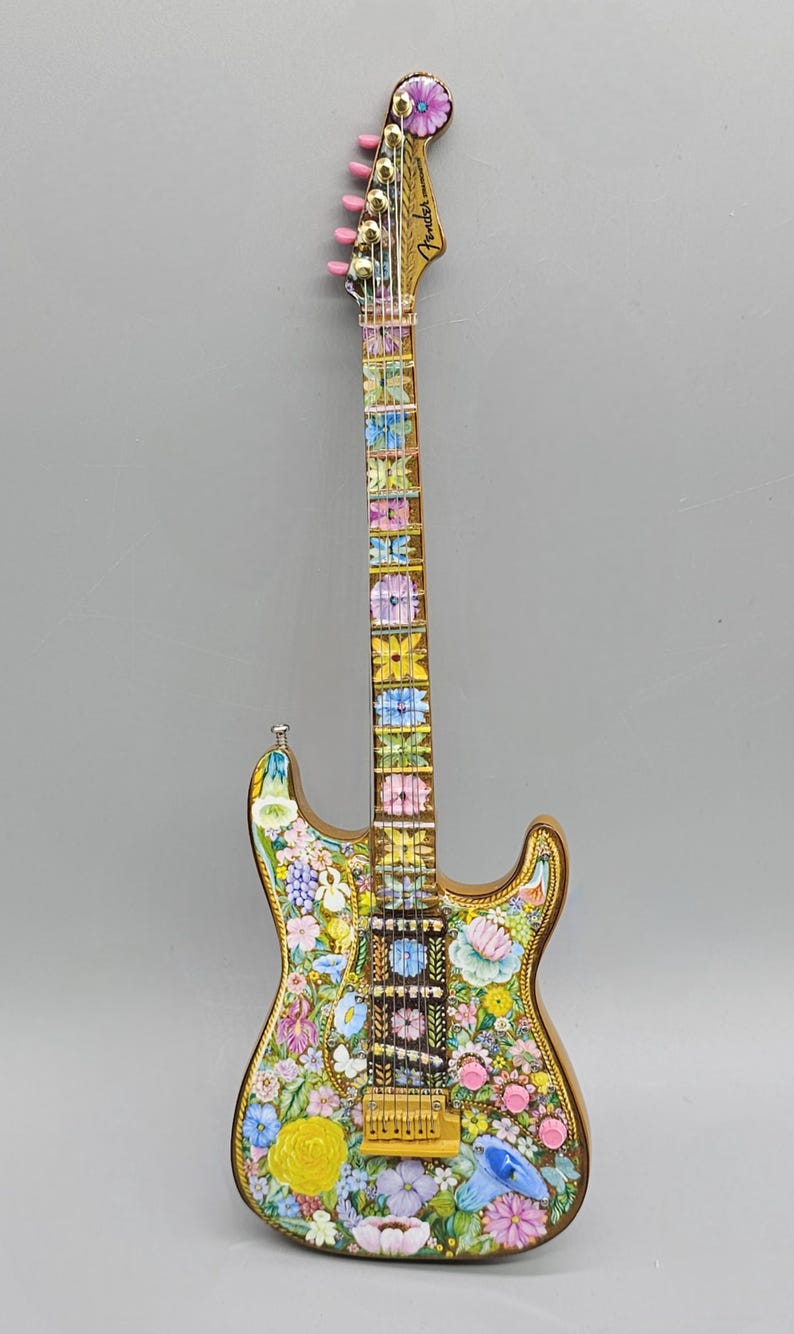 May include: A gold electric guitar with a floral design. The guitar has a pink flower on the headstock and the words "Fender Stratocaster" are visible. The body of the guitar is covered in a variety of flowers, including roses, daisies, and lilies.