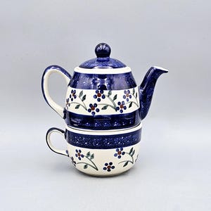 May include: A ceramic teapot and teacup set. The teapot has a lid and spout, and the teacup fits neatly underneath. Both pieces are white with blue stripes and a floral pattern of blue and brown berries.