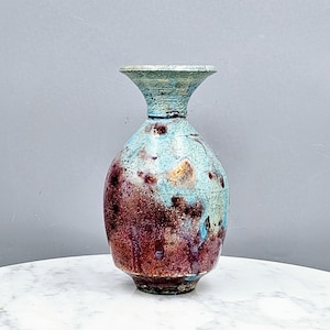 May include: A ceramic vase with a flared rim and a rounded body. The surface features a mottled glaze with blended shades of turquoise, deep red, and brown, creating a textured appearance across the exterior of the vessel from base to neck.