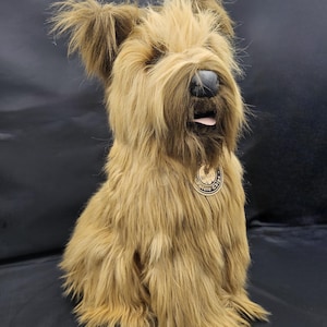 May include: A plush, seated dog with long, shaggy, light brown fur. The dog has a dark brown nose, a pink tongue, and a small tag around its neck. The dog's fur is a mix of light and dark brown, giving it a textured appearance.