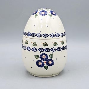 May include: An egg-shaped ceramic container with a lid, decorated with blue floral and leaf patterns. The off-white base is adorned with small blue dots. A decorative piece for the home.