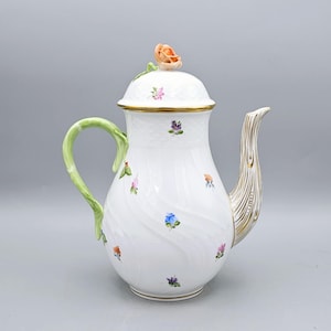 May include: White porcelain teapot with a lid topped with a peach-colored rose. The teapot features a green handle, a gold-trimmed spout, and is decorated with small, colorful floral designs. The teapot has a height of approximately 10 inches.