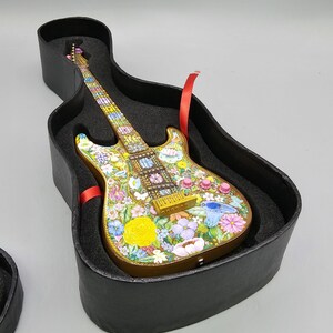 May include: A miniature electric guitar with a floral design painted on the body. The guitar is in a black case with a red ribbon.