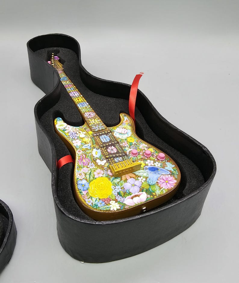 May include: A miniature electric guitar with a floral design painted on the body. The guitar is in a black case with a red ribbon.