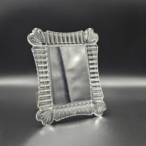 May include: A rectangular clear glass picture frame featuring a decorative border with vertical fluted ridges and fan-shaped corner accents. The frame has a smooth, reflective central panel designed to hold a photograph or display item securely.