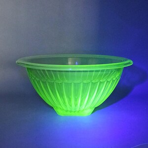May include: A translucent, vibrant neon green glass bowl featuring a wide, rounded rim and a series of vertical fluted ridges that extend from the base to a scalloped pattern just below the top edge, illuminated under ultraviolet light.