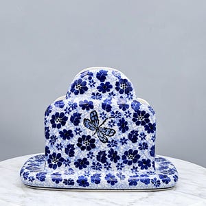 May include: A ceramic napkin holder featuring a white base with a dense pattern of small blue flowers and leaves. A single dragonfly illustration with blue wings and a black body is centered on the front panel of the arched, upright structure.