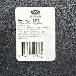 May include: A white label with black text and a barcode. The label reads "Item No. 12077 Flower Power Figurine GUITARMANIA&trade; &copy; 2013 United Way of Greater Cleveland FENDER&trade;, STRATOCASTER&trade; and the distinctive headstock and body design displayed here are the trademarks of Fender Musical Instruments Corporation. All rights reserved. Licensee: Westland Giftware, Inc. Made in China 7 48787 120"
