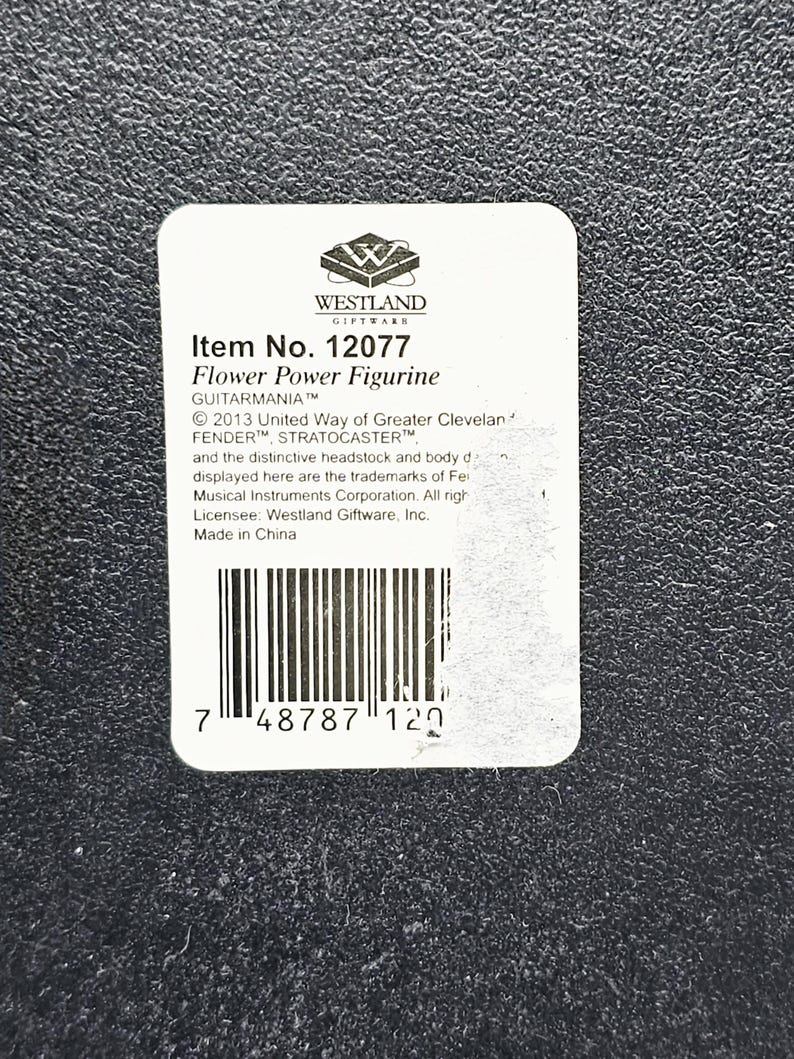 May include: A white label with black text and a barcode. The label reads "Item No. 12077 Flower Power Figurine GUITARMANIA&trade; &copy; 2013 United Way of Greater Cleveland FENDER&trade;, STRATOCASTER&trade; and the distinctive headstock and body design displayed here are the trademarks of Fender Musical Instruments Corporation. All rights reserved. Licensee: Westland Giftware, Inc. Made in China 7 48787 120"