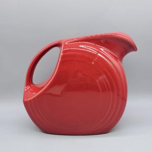 May include: A vibrant red ceramic pitcher with a curved handle and a spout. The pitcher has a rounded body with vertical ridges, and it sits on a solid base. The glossy finish reflects light, highlighting its smooth, curved design. A classic kitchenware item.