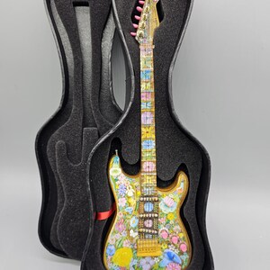 May include: A miniature electric guitar with a floral design in shades of pink, yellow, blue, and green. The guitar is in a black hard case.