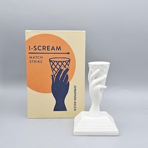 May include: A white ceramic candle holder in the shape of a hand holding an ice cream cone. The candle holder sits on a square base. Beside it is a matchbox with the words "I-SCREAM MATCH STRIKE" and a graphic of a hand holding an ice cream cone.