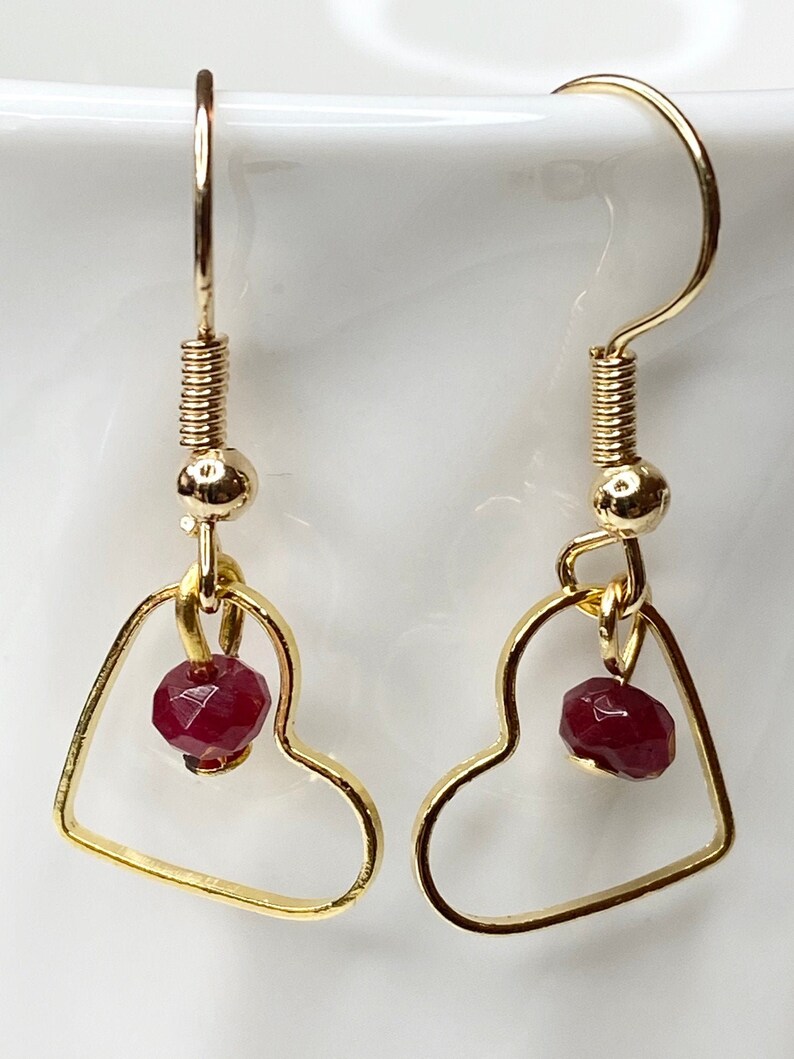 K&ouml;nnte beinhalten: Goldfarbene herzf&ouml;rmige Ohrringe mit einem kleinen, facettierten, roten Edelstein, der in jedem Herz h&auml;ngt. Die Ohrringe haben ein einfaches Haken-Design und sind vor einem wei&szlig;en Hintergrund platziert.