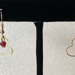 K&ouml;nnte beinhalten: Ein Paar goldfarbene herzf&ouml;rmige Ohrringe. Jeder Ohrring hat einen kleinen, runden, roten Edelstein, der innerhalb der Herzform h&auml;ngt. Die Ohrringe haben ein einfaches Haken-Design f&uuml;r durchbohrte Ohren.