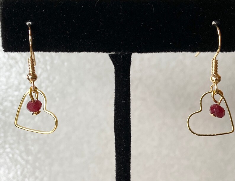 K&ouml;nnte beinhalten: Ein Paar goldfarbene herzf&ouml;rmige Ohrringe. Jeder Ohrring hat einen kleinen, runden, roten Edelstein, der innerhalb der Herzform h&auml;ngt. Die Ohrringe haben ein einfaches Haken-Design f&uuml;r durchbohrte Ohren.