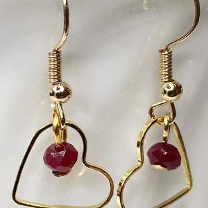 K&ouml;nnte beinhalten: Ein Paar goldfarbene herzf&ouml;rmige Ohrringe. Jeder Ohrring hat eine kleine, facettierte, dunkelrote Perle, die in dem Herzrahmen h&auml;ngt. Die Ohrringe haben ein schlichtes, elegantes Design mit einem Hakenverschluss.