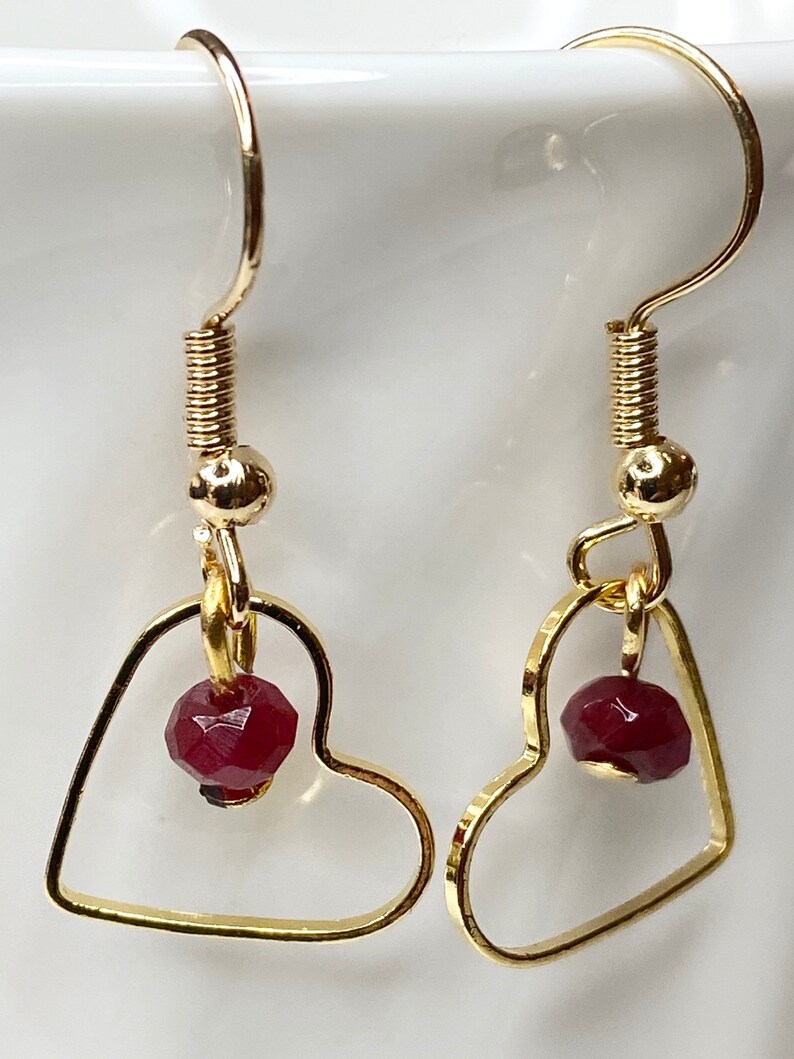 K&ouml;nnte beinhalten: Ein Paar goldfarbene herzf&ouml;rmige Ohrringe. Jeder Ohrring hat eine kleine, facettierte, dunkelrote Perle, die in dem Herzrahmen h&auml;ngt. Die Ohrringe haben ein schlichtes, elegantes Design mit einem Hakenverschluss.