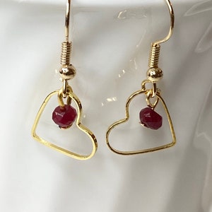 K&ouml;nnte beinhalten: Ein Paar goldfarbene herzf&ouml;rmige Ohrringe. Jeder Ohrring hat einen kleinen, facettierten, roten Edelstein, der in dem Herzrahmen h&auml;ngt. Die Ohrringe haben ein einfaches Haken-Design.