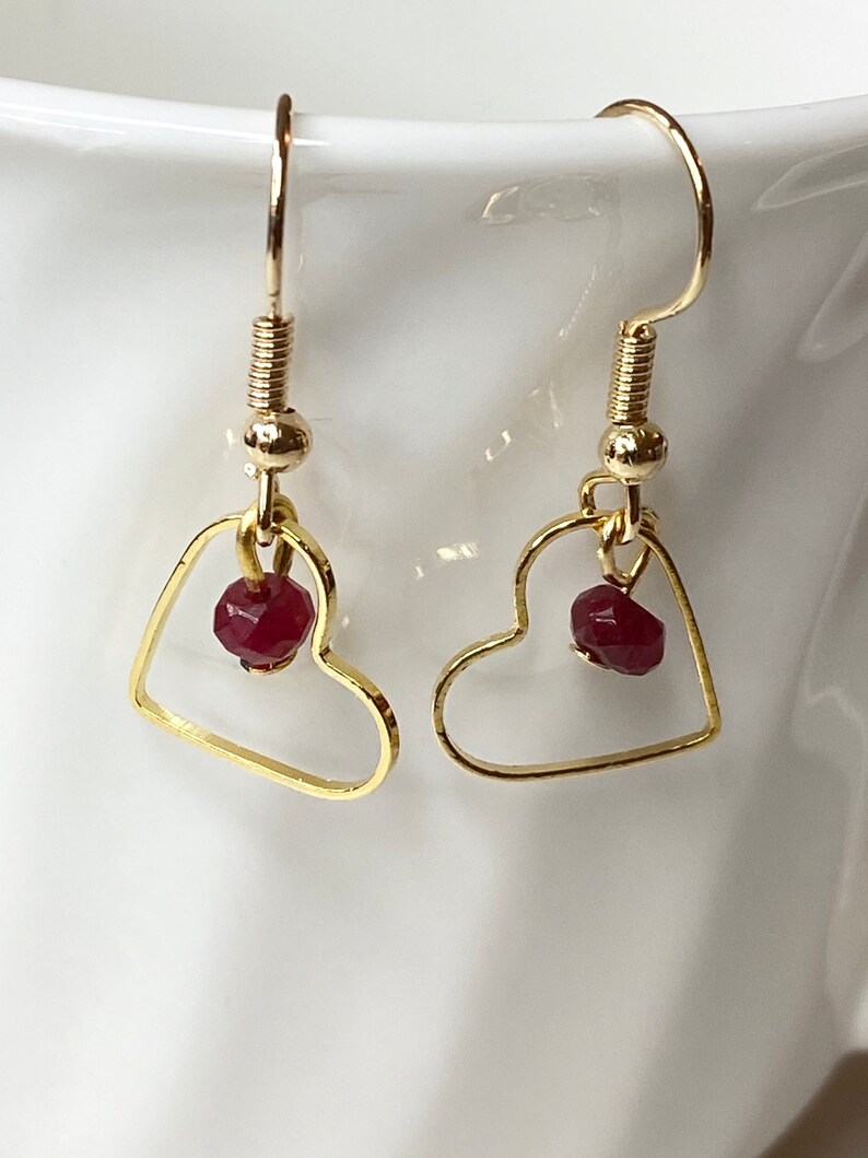 K&ouml;nnte beinhalten: Ein Paar goldfarbene herzf&ouml;rmige Ohrringe. Jeder Ohrring hat einen kleinen, facettierten, roten Edelstein, der in dem Herzrahmen h&auml;ngt. Die Ohrringe haben ein einfaches Haken-Design.