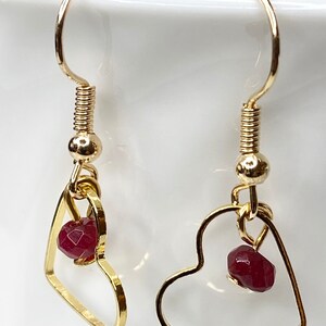 K&ouml;nnte beinhalten: Goldfarbene herzf&ouml;rmige Ohrringe. Jeder Ohrring hat einen kleinen, facettierten, tiefroten Edelstein, der in dem Herzrahmen h&auml;ngt. Die Ohrringe haben ein schlichtes, elegantes Design mit Hakenverschluss.