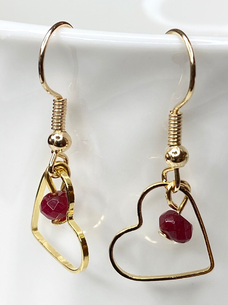 K&ouml;nnte beinhalten: Goldfarbene herzf&ouml;rmige Ohrringe. Jeder Ohrring hat einen kleinen, facettierten, tiefroten Edelstein, der in dem Herzrahmen h&auml;ngt. Die Ohrringe haben ein schlichtes, elegantes Design mit Hakenverschluss.