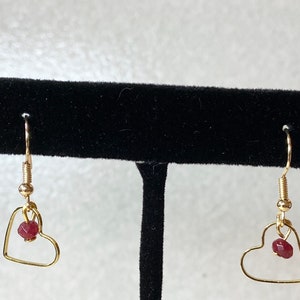 K&ouml;nnte beinhalten: Ein Paar goldfarbene herzf&ouml;rmige Ohrringe. Jeder Ohrring hat einen kleinen, roten Edelstein, der in dem Herzrahmen h&auml;ngt. Die Ohrringe haben ein schlichtes, elegantes Design mit einem Hakenverschluss.