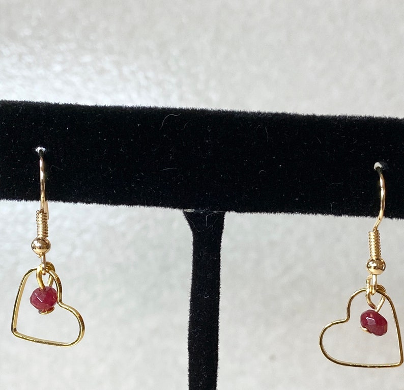 K&ouml;nnte beinhalten: Ein Paar goldfarbene herzf&ouml;rmige Ohrringe. Jeder Ohrring hat einen kleinen, roten Edelstein, der in dem Herzrahmen h&auml;ngt. Die Ohrringe haben ein schlichtes, elegantes Design mit einem Hakenverschluss.