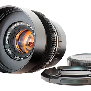 以下が含まれることがあります： ブラックのOlympus OM-System Zuiko f=50mm 1:1.8カメラレンズ。レンズフードが装着されており、ギザギザのフォーカスリングと透明なガラスエレメントが特徴です。黒いフロントレンズキャップと、FX MOUNTと記されたリアレンズキャップが付属しています。
