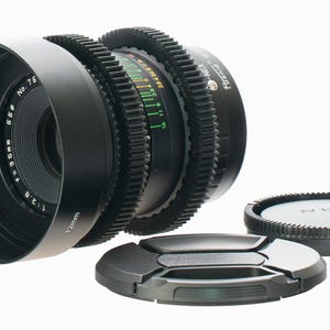 May include: A black camera lens with a lens hood and two lens caps. The lens has a 72mm diameter and f/2.8 aperture. The lens hood is labeled "No. 76". The lens caps are black with the brand name "NEX" on one.