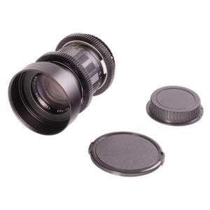 Puede incluir: Un objetivo de cámara negro con el texto "TEL AUTO 1:2.8" y dos tapas de objetivo. El objetivo tiene un anillo de enfoque estriado y marcas de medición. Las tapas son negras y redondas.