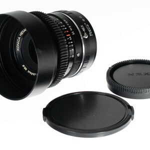 May include: A black Konica Hexanon 35mm camera lens with a lens hood and two lens caps. The lens has a focus ring and aperture settings. The lens caps are black and round. The lens is labeled "Konica Hexanon Japan 55".