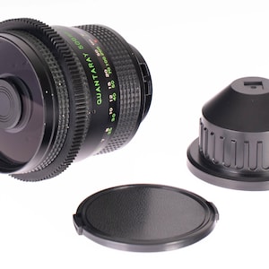 May include: A black Quantaray 500mm camera lens with a lens cap and a lens hood. The lens has green and white markings and a ridged focus ring. The lens cap is round and textured. The lens hood is black and cylindrical.