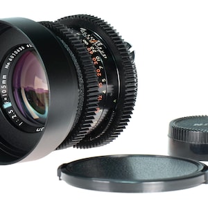 May include: A black camera lens with a lens hood, focus rings, and a lens cap. The lens has the text "f=105mm 1:2.5" and "No. 6010636". A second lens cap is also present. The lens is for photography.