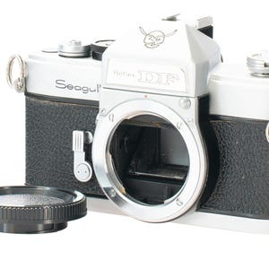 May include: A silver and black Seagull Reflex DF film camera body with a textured black grip, a silver top plate, and a circular lens mount. A separate black circular lens cap sits in front of the camera body on a white surface.