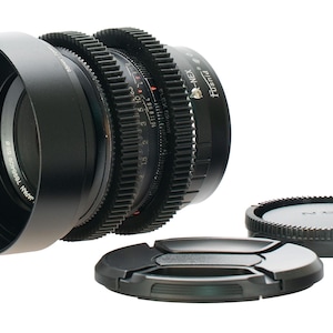 May include: A black camera lens with a lens hood, focus rings, and two lens caps. The lens has a series of gears and markings. The lens caps are black and round. The lens is designed for a NEX camera.