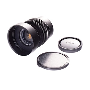 May include: A black camera lens with a lens hood and two lens caps. The lens has a ridged focus ring and markings. One cap is labeled "NEX". The lens is a 37mm lens.