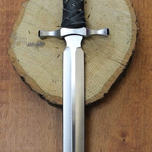May include: A silver dagger with a black leather handle and a woven design. The dagger is on a wooden surface.