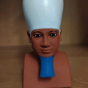 Peut inclure: Buste d'un pharaon égyptien avec une couronne blanche, une peau brune et un collier bleu. Les yeux sont détaillés avec un eye-liner noir. Le buste est de couleur brun rougeâtre.