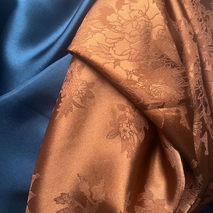 Op de afbeelding: Een close-up van twee stoffen, een in diepblauw en de andere in koperkleur met een bloemenpatroon. De koperkleurige stof heeft een subtiele glans en een reliëf bloemenpatroon.