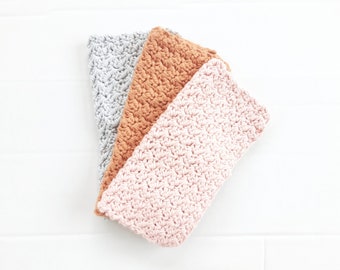 Puede incluir: Tres paños de cocina de ganchillo en diferentes colores: gris, naranja y rosa. Los paños de cocina están doblados y apilados uno encima del otro.