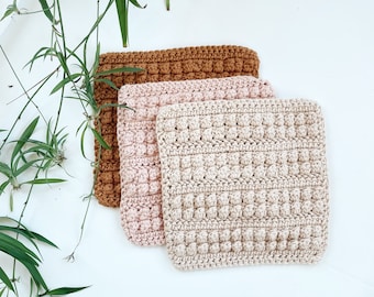 Puede incluir: Tres paños de cocina de ganchillo en marrón, rosa y beige. Los paños de cocina están apilados uno encima del otro.