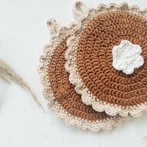 May include: Two brown and beige crocheted potholders with scalloped edges and hanging loops. A small white crocheted flower is placed on one of the potholders. The potholders are handmade and have a rustic, cozy aesthetic.