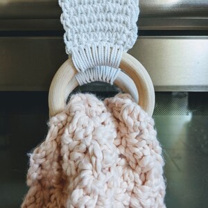 Shows the towel hanger in use. one wooden ring is behind the handle of the oven and one wooden ring is in front of it. A blush pink towel is placed inside both of the rings. The towel rests inside of the wooden rings.