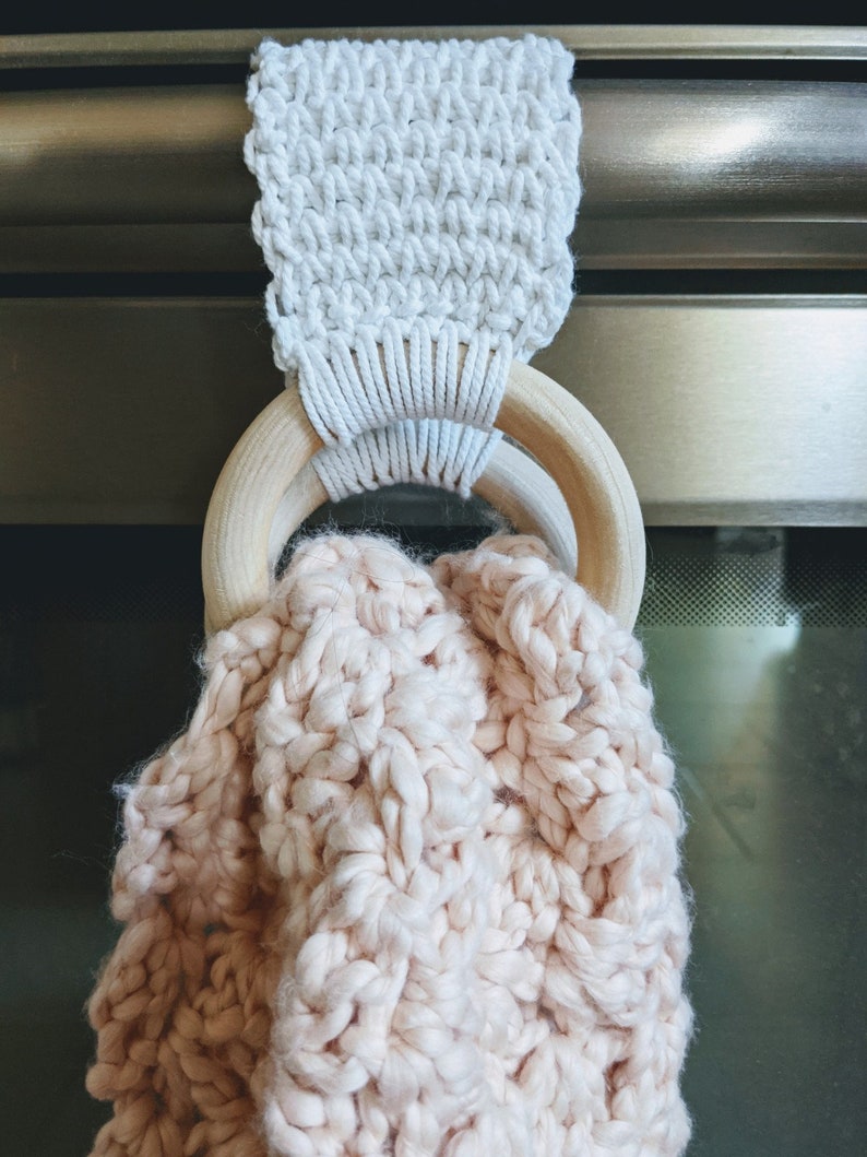 Shows the towel hanger in use. one wooden ring is behind the handle of the oven and one wooden ring is in front of it. A blush pink towel is placed inside both of the rings. The towel rests inside of the wooden rings.