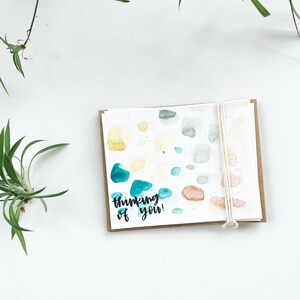 May include: A card with a watercolor design of colorful dots and the text "thinking of you!" on a white background.