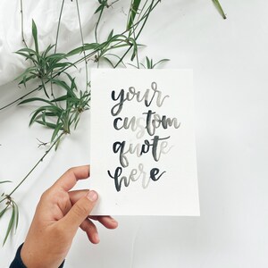May include: A watercolor card with the words "your custom quote here" in a gradient of gray and black calligraphy. The card is held in a hand, with green plants and white fabric in the background.