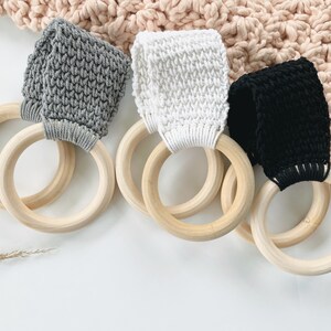 Puede incluir: Tres anillos de dentici&oacute;n de madera con correas de crochet de diferentes colores. Los anillos son de madera natural y las correas son grises, blancas y negras.